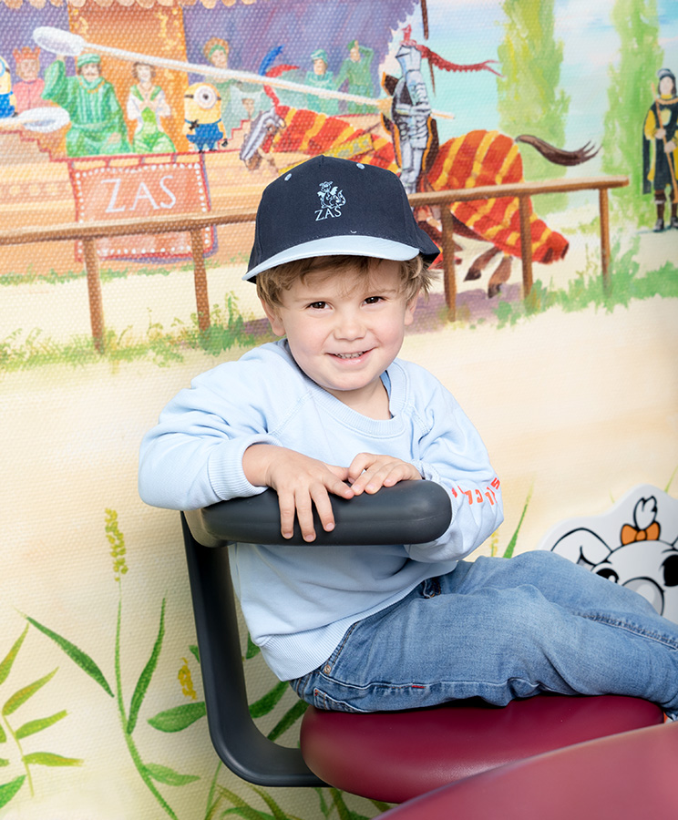 Kleiner Junge mit Cappie lächelt ein kleiner Junge mit Cappie sitzt auf einem Hocker vor einer bemalten Wand mit Märchenbildern und lächelt