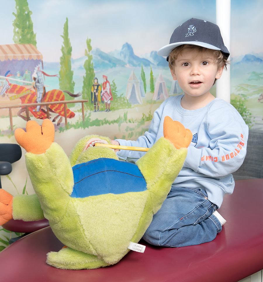 Kleiner Junge mit Cappie Kleiner Junge mit Cappie auf dem Kopf kniet auf im Kinderbehandlungsraum der Praxis und putzt einem Stofftierdrachen die Zähne mit einer riesigen Zahhbürste